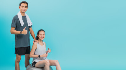 Wall Mural - Asian beautiful happy woman holding water bottle and sitting on fit ball with boyfriend after exercise isolated on blue color background.Concept of slim and healthy girl workout.