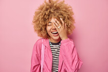 Joyful Young European Woman Makes Face Palm And Smiles Broadly Hears Funny Joke Keeps Eyes Closed Dressed In Windbreaker And Striped Jumper Isolated Over Pink Background. Happy Emotions Concept