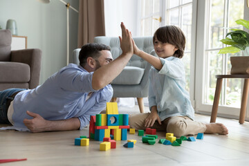 Happy proud dad and cheerful little son boy clapping hands, giving high five over toy castle, tower of wooden construction blocks on clean warm floor, celebrating model completing