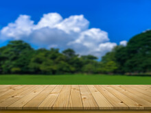 Empty Perspective Brown Plank Wooden Board Mock Up Display As Shelf Or Table With Blurred Background Sunshine Blue Sky And White Clouds On The Green Tree And Grass Field.