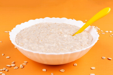 Wall Mural - Oatmeal porridge for the baby in a white bowl (plate) and grits on a yellow background close-up. The first complementary food, baby nutrition. Healthy breakfast. Side view.