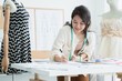 © Pruksachat - Asian beautiful woman fashion designer or dressmaker with tape measure on neck, take note of measured size on paper, new dress collection on a mannequin, working with happiness at workplace studio