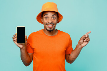 Young man of African American ethnicity wear orange t-shirt hat hold use mobile cell phone with blank screen workspace point finger aside on area isolated on plain pastel light blue cyan background.