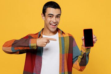 Young satisfied happy fun middle eastern man 20s wear casual shirt white t-shirt hold use point index finger on mobile cell phone with blank screen workspace area isolated on plain yellow background.