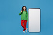 © ViDi Studio - Full body young woman of African American ethnicity wear green shirt near big huge blank screen mobile cell phone with workspace copy space mockup area show thumb up isolated on plain blue background.