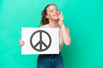 Wall Mural - Young caucasian woman isolated on green background holding a placard with peace symbol and shouting