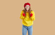 Young girl in yellow sweatshirt, red hat and gloves and jeans making a wish on beige background. She is smiling closing her eyes and holding her hands in fists. Desire, dream, fulfillment concept.