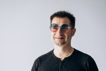 Wall Mural - Handsome man in a black t-shirt and round glasses stands and looks confidently at the camera and smiles.