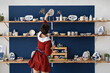 © DimaBerlin - Back view of ceramist girl tidies up the shelves with handcraft ceramics at her studio. Pottery studio owner putting plate at right place. Small business and female artist entrepreneur concept