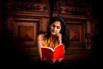 Young beautiful East Indian American college female student sitting by vintage style door way, tilting head, hand touching chin, looking down, reading red book..