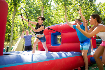 Emotional woman tied with rope to her friend competing to collect hoops on inflatable playground on sunny summer day..