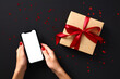 © photoguns - Female hands holding smartphone mockup with blank screen over black background with craft paper gift box wrapped red ribbon bow. Black Friday sale concept.