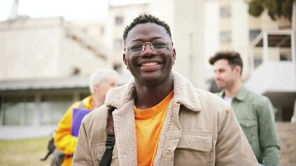 Wall Mural - Young african american student with googles smiling and looking at camera standing at the high school, at the back ground a group of teenagers walking and laughing. Education concept. High quality 4k