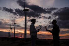 Engineers Discussing With Each Other Holding Laptop At Wind Farm On Sunset