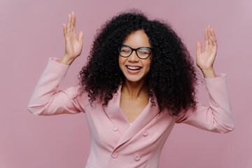 Wall Mural - Overjoyed emotional female entrepreneur raises hands, laughs sincerely, wears spectacles and formal jacket, works in business sphere, poses indoor against purple background. Happy emotions concept