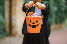 Little Cute Girl In Witch Costume Holding Jack-o-lantern Pumpkin Bucket With Candies And Sweets. Kid Trick Or Treating In Halloween Holiday.