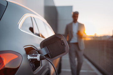 Close up of electric car charging, businessman standing in background and waiting.