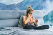 Bedroom interior. Young caucasian mom sitting cross-legged on bed, breastfeeding her infant baby boy, and talking via smartphone. Full body shot. . High quality photo