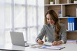© ArLawKa - Smiling businesswoman working in office with money chart document and holding pen using laptop sitting on chair at studio in office accounting concept