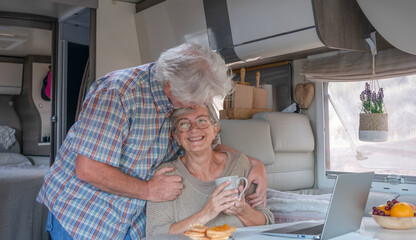 Wall Mural - Elderly white-haired man kissing his wife in travel vacation inside a camper van enjoying breakfast together. Happy caucasian senior couple in van life while browsing on laptop.