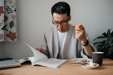 Adult asian man wearing glasses reading book or magazine while having breakfast. Leisure morning time.