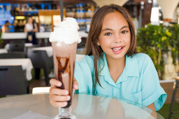 Beautiful child kid girl eating a chocolate shake in a restaurant. Cold summer desserts for kids. Happy authentic childhood lifestyle.