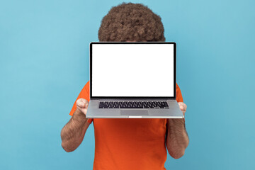 Unknown anonymous man with Afro hairstyle wearing orange T-shirt hiding his face with laptop with white empty display, copy space for promotional text. Indoor studio shot isolated on blue background.
