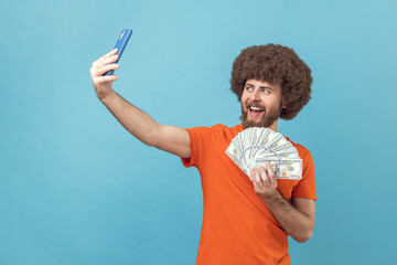 Portrait of positive optimistic man with Afro hairstyle wearing orange T-shirt having video call or livestream, showing dollar banknotes to followers. Indoor studio shot isolated on blue background.
