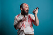 Spooky halloween monster holding smartphone in studio, using mobile phone app while being aggressive and scary. Frightening undead zombie with bloody wounds, cruel corpse with telephone.