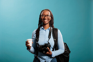 Wall Mural - Portrait shot of happy african american professional photographer with camera device smiling on blue background. Young adult woman with photography hobby having cup of coffee. Studio shot.