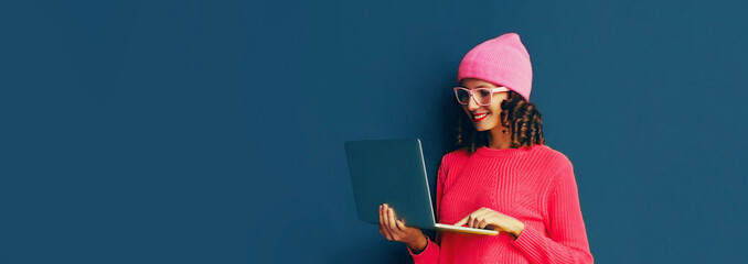 Portrait of stylish modern young woman working with laptop wearing pink colorful clothes on dark blue background, banner blank copy space for advertising text