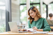 © makibestphoto - Happy asian business woman holding notebook paper and working with laptop in green sweater sitting at office desk.