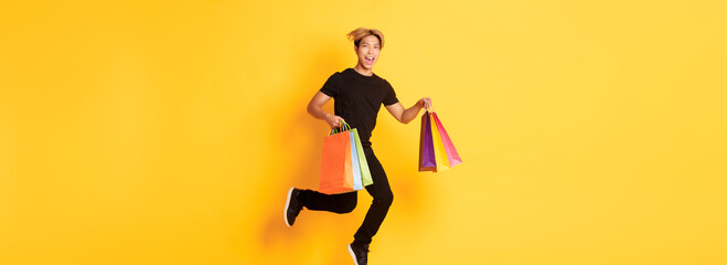 Full-length of happy attractive asian man jumping from happiness and carry shopping bags, standing yellow background