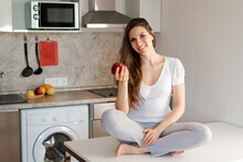 Young Slim Woman Standing In Kitchen On Table, Holding Red Apples, Looking At The Camera, Smiling Cheerful In Light Casual Clothes. Healthy Lifestyle Concept