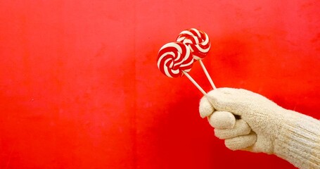 Against a rich red background, a hand in a beige winter glove holds two striped lollipops. Lollipops are spirally twisted red and white Sweets for Christmas and New Year holidays.