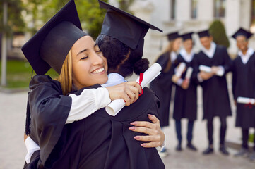 Wall Mural - Positive women graduates in student gown hug each other after receiving university or college diploma and rejoice at completion of their education standing under open sky. Selective background