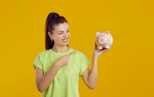 Smiling Woman Points To Piggy Bank In Her Hand, Hinting At Favorable Offer From Bank. Happy Caucasian Casual Woman Holding Piggy Bank In Form Of Pink Pig. Isolated On Vivid Yellow Background. Banner.