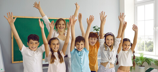 Portrait of funny group of elementary school classmates and their female teacher having fun in classroom. Smiling schoolchildren and teacher stand with raised hands. Panoramic web banner.