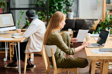 Poster - Young businesswoman with financial document looking at laptop screen while sitting by workplace and analyzing online information