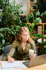 Poster - Young creative woman with long blond hair having coffee and making notes in document while analyzing online data on laptop screen