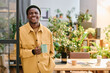 © pressmaster - Young smiling African American businessman with cup of tea or coffee looking at camera while standing against his workplace in office