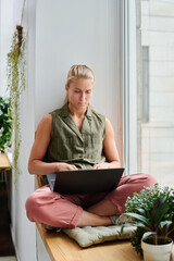 Poster - Young serious blond woman in casualwear with laptop on her crossed legs typing on keypad while searching for online information