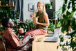 © pressmaster - Young smiling female office manager eating wok and talking to African American male colleague sitting by workplace among green plants