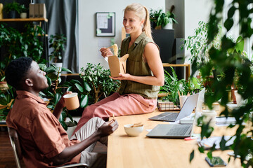 Aufkleber - Young smiling female office manager eating wok and talking to African American male colleague sitting by workplace among green plants