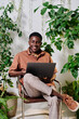 © pressmaster - Young smiling employee in casualwear sitting on chair with laptop on his knees and organizing work smong green plants in office
