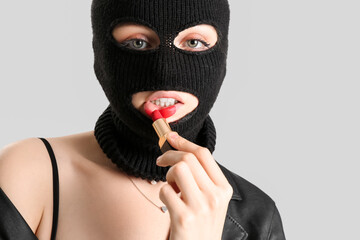 Wall Mural - Young woman in balaclava applying red lipstick against light background