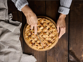 Canvas Print - A woman cuts a baked round pie with apples with a knife on a wooden table