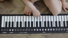 Baby Hands Playing Pushing Keys Electronic Piano On Wooden Floor Indoors High Angle View. Unrecognizable Toddler Sit On Floor Living Room Playing Soft Synthesizer White Black Piano Keys.