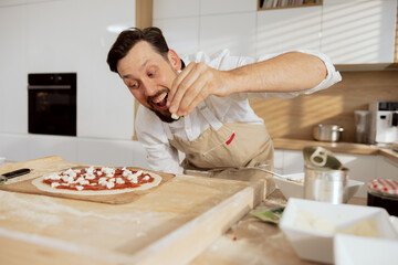 Delighted curious father husband dad wearing apron looking at homemade pizza applying feta on tomato sauce. Standing at wooden table in modern kitchen. Preparing lunch for children kids son daughter.