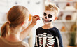 © JenkoAtaman - Festive makeup for Halloween. Woman doing skeleton make-up for boy in costume while preparing holiday
